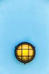 Ship yellow bulkhead light (or deck lamp) surrounded by a metal rusted frame fixed to a painted light blue pastel color wooden wall.