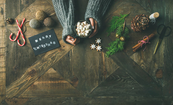 Christmas or New Year holiday preparation background. Flat-lay of greeting card, glittering toys, woman's hands in grey sweater holding mug of hot chocolate, cinnamon, candy cane, scissors over