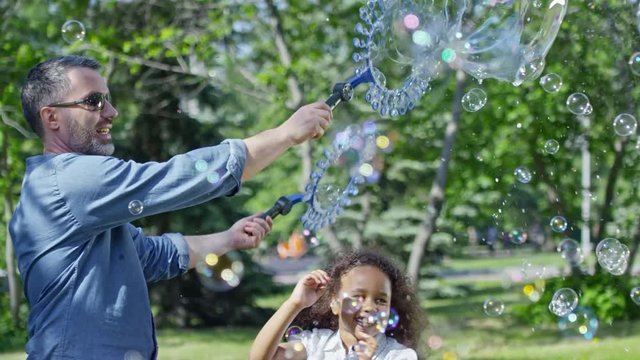 Slowmo of professional bubble artist blowing lots of small and large bubbles in the park while performing show for kids; little boys and girl jumping and catching bubbles