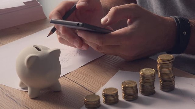 Man calculating home budget figures on mobile phone while sitting at the desk with stacked coins and piggy coin bank.
