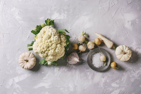 Variety of white vegetables raw organic cauliflower, pumpkins, garlic, parsnip and radish with plate of sea salt over gray texture background. Top view with space. Healthy eating concept. Toned image