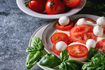 Italian caprese salad with sliced tomatoes, mozzarella cheese, basil, olive oil. Served in ceramic plate over gray texture background. Close up
