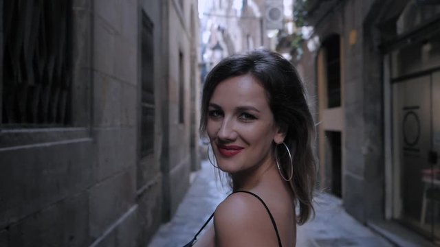 Gorgeous inviting woman in black dress with passionate red lips turns and looks to the camera. Natural beauty, female portrait, seductive look. Traveling Barcelona street. Slow motion.