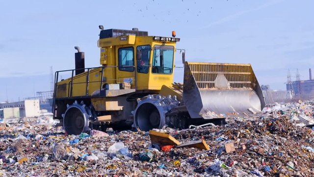A landfill compactor moves with a raised blade at a city landfill. Waste, garbage, dump, rubbish 4K.