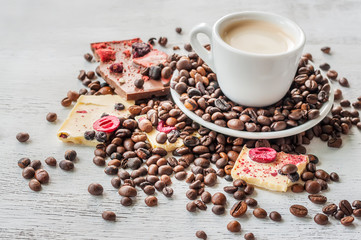 Coffee in a cup and scattered coffee beans around and pieces of white chocolate with fruit on a light wooden background.
