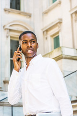 Young African American Man working in New York, wearing white shirt, holding laptop computer, standing on stairs of vintage office building doorway, looking away, talking on cell phone..