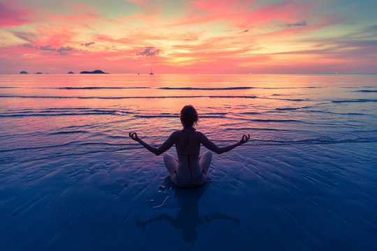Young woman practicing yoga sitting on the sea beach during an amazing sunset.