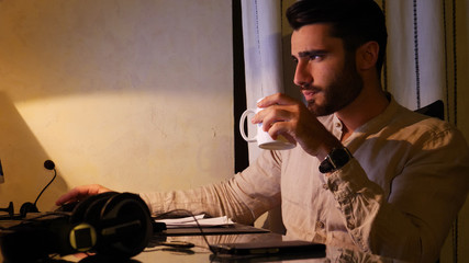 Attractive young businessman sitting at desk using computer at home and drinking coffee or tea