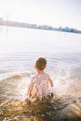 boy child playing makes splashes, beats hands on the water in the river at sunset