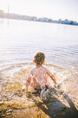 boy child playing makes splashes, beats hands on the water in the river at sunset