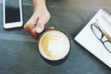 coffee cup in hand business woman ready to drink in the morning of work. Office desk table with book notepad, phone, equipment supplies in the work. Top view with copy space. 