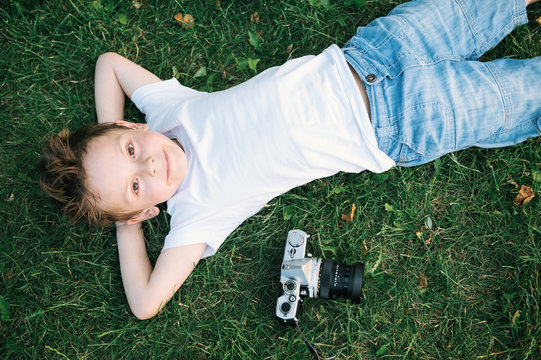 happy smiling little boy lying on the green grass near vintage film camera