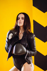 Obraz na płótnie Canvas Seductive woman in leather jacket poses in boxing gloves before a yellow wall