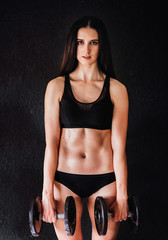 Fototapeta na wymiar Woman in black fitness suit stands with barbells before a black wall