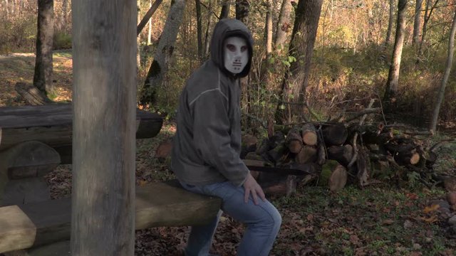 Man with scary Halloween mask and machete sitting in  park


