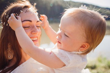 Fototapeta premium Mother in white dress plays with her charming daughter in white gawn before a lake