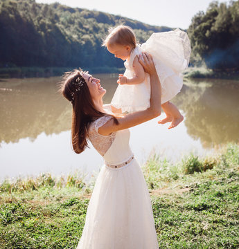 Mother in white dress plays with her charming daughter in white gawn before a lake