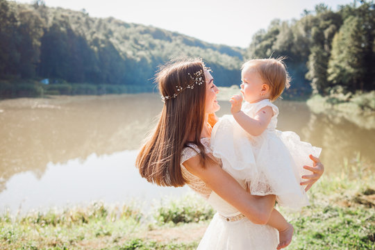 Mother in white dress plays with her charming daughter in white gawn before a lake