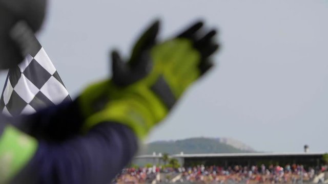 Man holding and waving Checkered race flag in slow motion at finish line on a raceway. Hands of unidentified person applauding.Victory, achievement, success and sport concept.