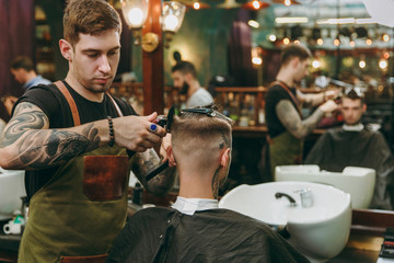 A man getting trendy haircut at barber shop. Male hairstylist in tattoos serving client.