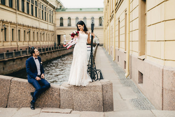 Handsome bridegroom sits on bridge looks at his beautiful bride who stands up, says compliments to her, enjoy togetherness, have unforgettable moments together, Romantic people and wedding concept
