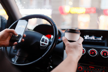 Stylish driver with a smartphone in hand and paper cup of hot coffee in the driver's seat. The concept of inattention at the wheel, rest, coffee break to cheer.