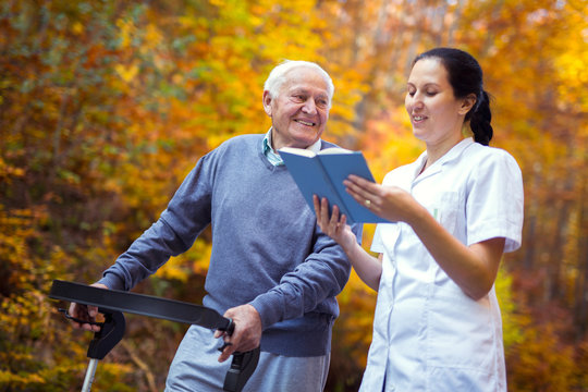 Smiling nurse reading book to senior man that uses walker with caregiver outdoor