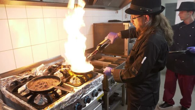 Two male chef adding wine in a pan full of meat and veggie mix in restaurant kitchen cooking a tasty flambe 