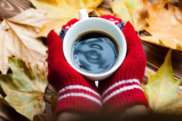 Hands in red knitted gloves holding a hot cup of coffee against yellow leaves background. Concept...