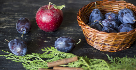 Purple organic plums, apple and autumn decoration on dark wooden table.