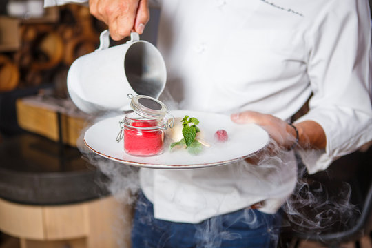 Hands chef holding a white plate with ice cream dessert and pouring from the jug with dry
