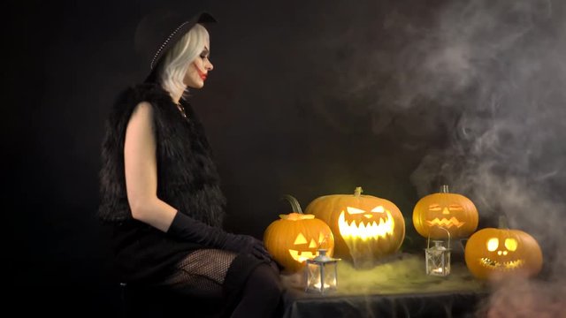 Female sorceress with gray hair scares, conjures, casts a spell. Scary beautiful girl witch celebrates halloween with funny glowing burning pumpkins in smoke. Gates of hell in fire