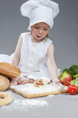 Cooking Ideas and Concepts. Portrait of Little Caucasian Girl in Cook Hat and Lips in Flour Sitting in Front of Fresh Buns and Fruits.Against Gray.