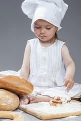 Cooking Ideas and Concepts. Portrait of Little Caucasian Girl in Cook Hat and Lips in Flour Sitting in Front of Fresh Buns. Against Gray.