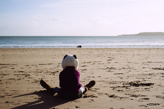 A little girl in a panda hat sitting on a beach looking at the sea.