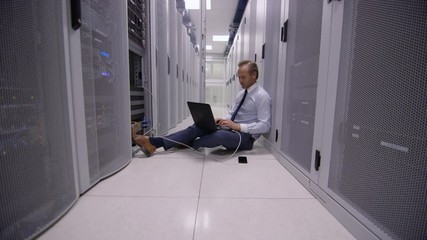 Stressed computer engineer sitting on floor and working on laptop, checking the servers in a data center.