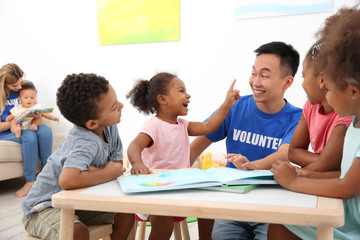 Young male volunteer reading book with little children in light room. Volunteering abroad concept