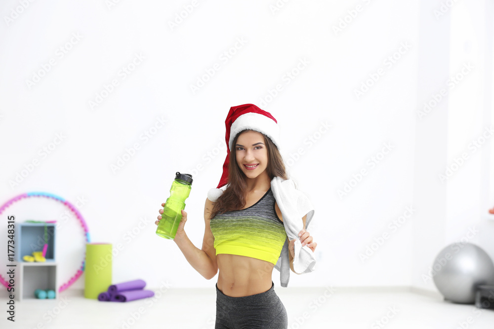 Wall mural beautiful young woman in santa claus hat with bottle of water at gym