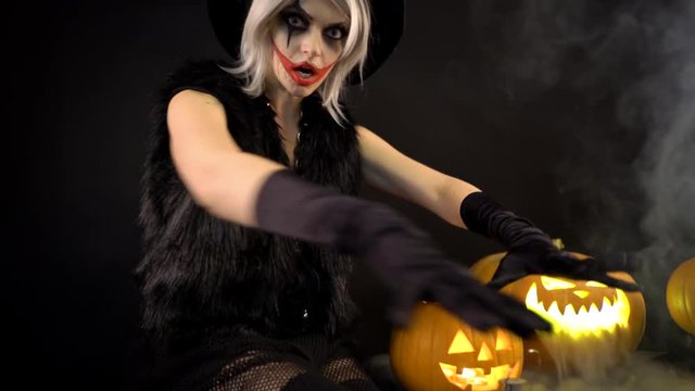 Female sorceress with gray hair scares, conjures, casts a spell. Scary beautiful girl witch celebrates halloween with funny glowing burning pumpkins in smoke. Gates of hell in fire