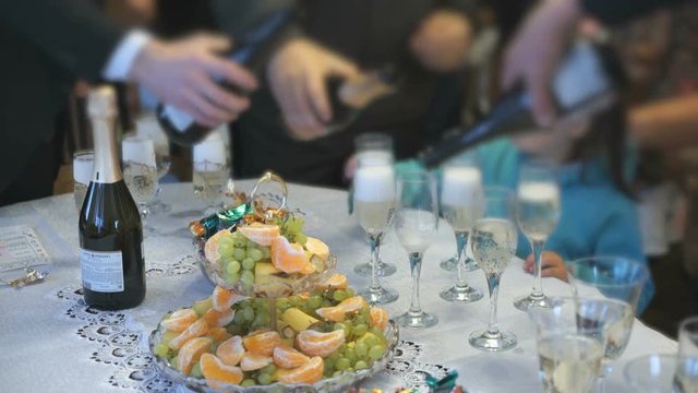 Wedding feast - unidentified men in formal clothes pour sparkling wine into glass on the background of table with food - fruits, candy, snacks
