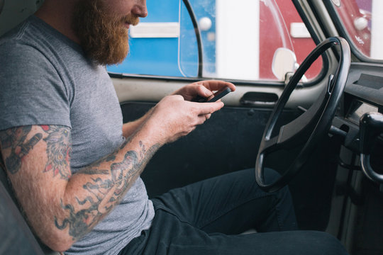 Young bearded man with tattoos texting with his cell phone in a vintage car