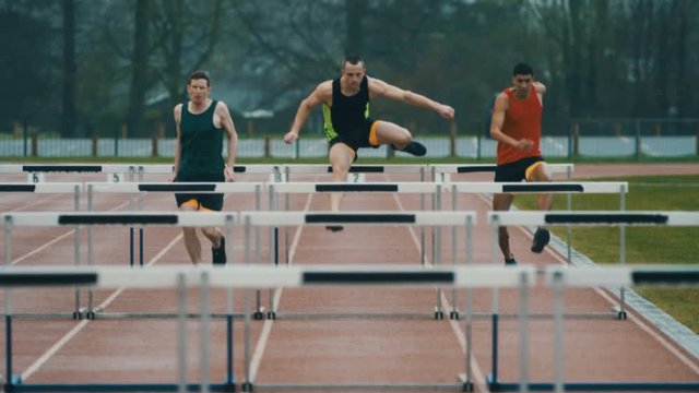  Competitive athletes running & jumping over hurdles at athletics track