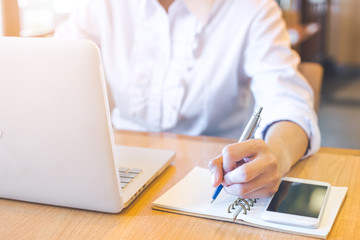 A business woman hand working on a laptop computer and writing on notepad with a pen on her office desk.