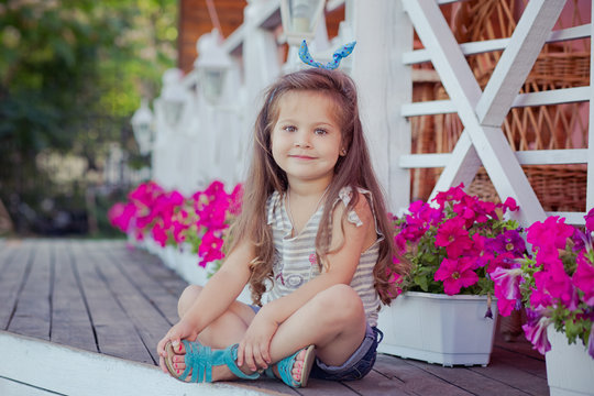 Stylish beautifull cute baby girl with brunette hair posing on wooden garden full of flowers wearing tiny jeans shirts and airy skivy underwaist and blue sandals