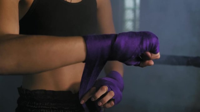 Closeup view of female hands being wrapped for boxing in dark room with smoke. Young fit woman wrapping hands with purple boxing bandage. Shot in 4k