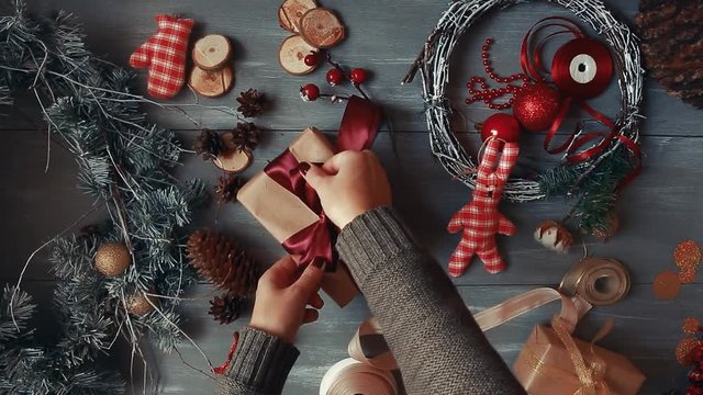 A top-down plan. Fully visible the table with the decorations. Female hands put and finalize Christmas gift wrapped in craftool paper on a wooden table. Bandaging tape and tied in a bow.