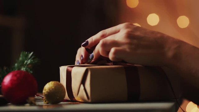 Close-up on the background of Christmas lights female hands open a gift from Kraft paper. The background is blurred in bokeh.