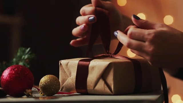 Close-up on the background of Christmas lights women's hands tying the ribbon on the gift of Kraft paper. The background is blurred in bokeh.