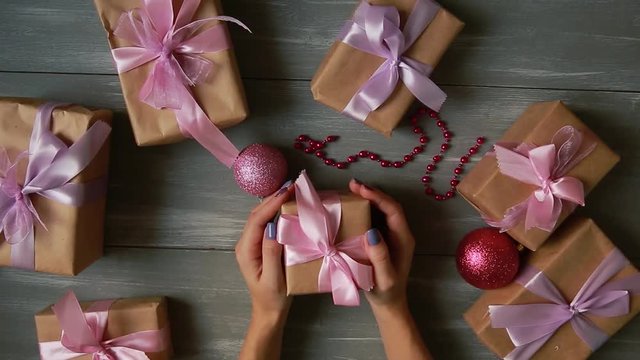 A top-down plan. Fully visible the table with the decorations. Female hands put and finalize Christmas gift wrapped in craftool paper on a wooden table. Bandaging tape and tied in a bow.