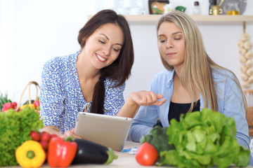 Two young happy women making online shopping for making menu by tablet computer. Friends cooking in the kitchen